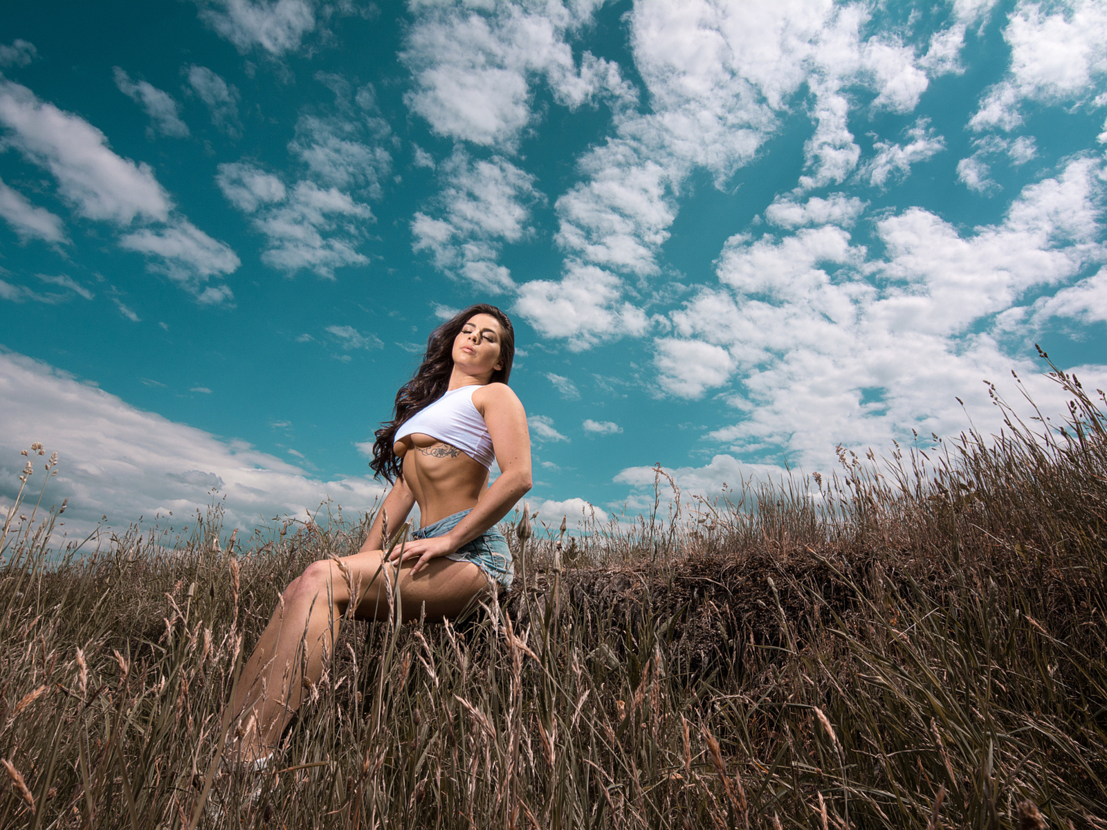 women, closed eyes, underboob, t-shirt, sitting, women outdoors, sky, clouds, tattoo, long hair, wavy hair, belly, brunette, boobs, jean shorts