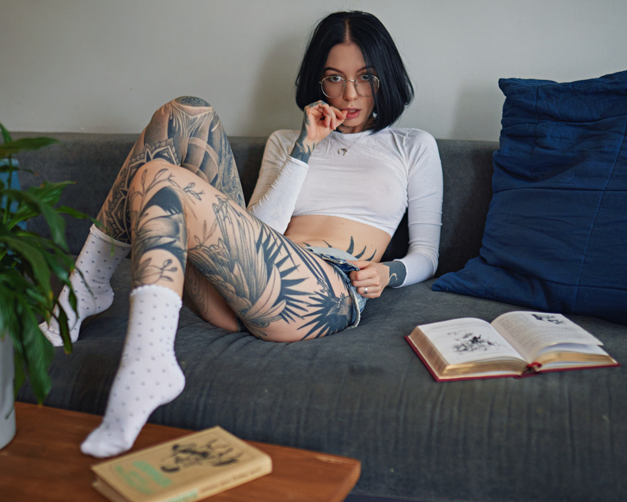 women, sergey fat, couch, tattoo, glasses, sitting, white socks, women indoors, books, plants, finger on lips, jean shorts, women with glasses, necklace, black hair, see-through clothing, nipples through clothing