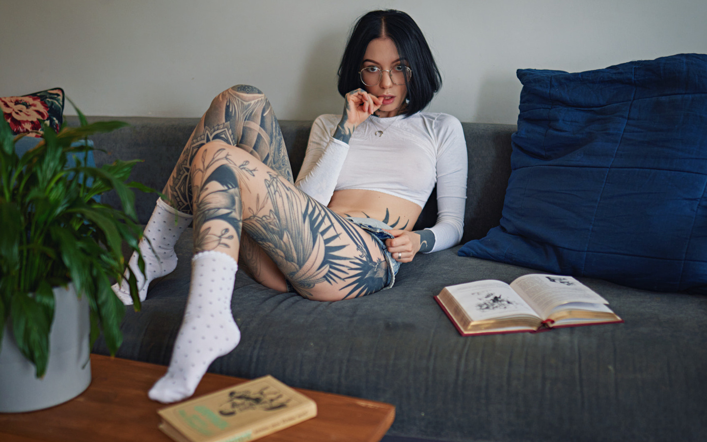 women, sergey fat, couch, tattoo, glasses, sitting, white socks, women indoors, books, plants, finger on lips, jean shorts, women with glasses, necklace, black hair, see-through clothing, nipples through clothing