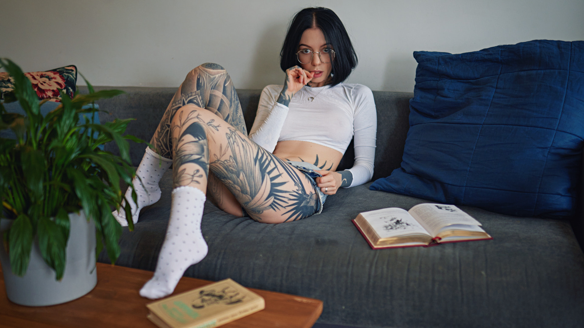 women, sergey fat, couch, tattoo, glasses, sitting, white socks, women indoors, books, plants, finger on lips, jean shorts, women with glasses, necklace, black hair, see-through clothing, nipples through clothing